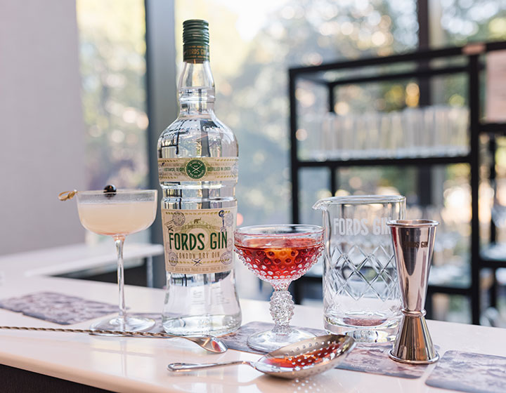 A Ford's Gin bottle on an elegant bar, set next to two cocktails, a shaker, and other accessories